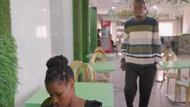 Shy couple meeting bright cafe, young woman seated at table scrolling phone, man in striped sweater walks over and places gentle hand on shoulder, soft smiles exchanged, modern bistro with green