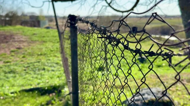 Rusty chain fence on garden plot under bare tree branches, leaning metal post and old pipe casting long shadow across fresh spring.