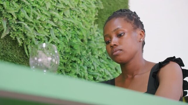 Woman leaning at green cafe table, studying glass reflection with calm expression, gathering resolve after missed rendezvous, indoor moss wall backdrop, warm natural light, subtle cinematic closeup