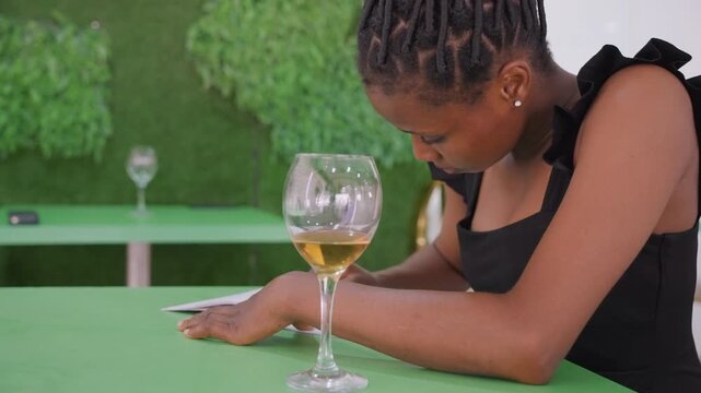 Black woman waiting at green table glass of wine by phone checking messages head bowed braided hair elegant black dress outdoor cafe hedge backdrop melancholic mood romantic disappointment ghosting