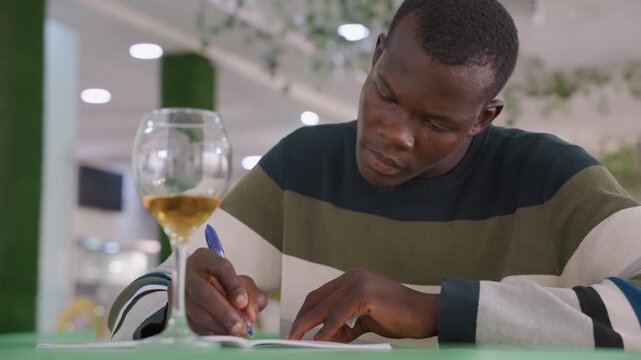 Man composing apology note at cafe, wine glass beside him, tense expression, striped sweater, low light, handwritten card, closeup of pen, intimate bistro setting, melancholic mood.