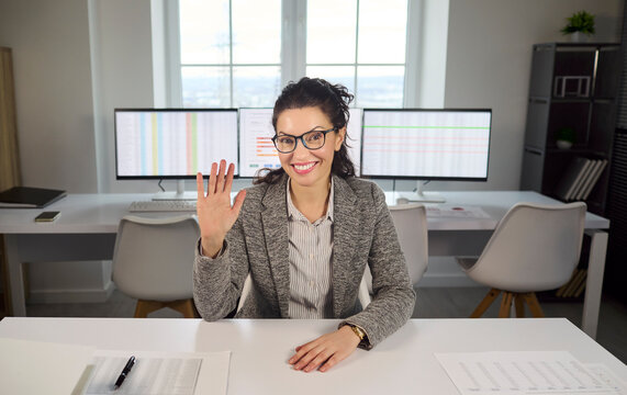 Woman businesswoman office video call online meeting. Smiling professional in glasses waving at desk with dual monitors, greeting clients. Concept of remote business communication online.