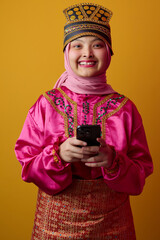 Naklejka premium Smiling Young Muslim Indonesian Asian Woman In Bright Traditional Costume And Hijab Holding Smartphone For Celebration