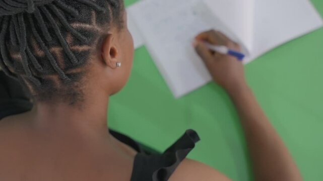 Black woman writes love note at table, braided hair and ribbon strap visible, pen gliding over paper, closeup overshoulder framing, green tabletop, soft natural light, intimate thoughtful mood,