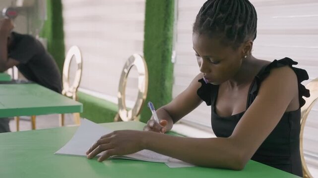Young woman writing love letter, table. Focused scene in bright bistro, green tablecloth, pages spread, pen moving across paper, quiet afternoon vibe, distant patron in background
