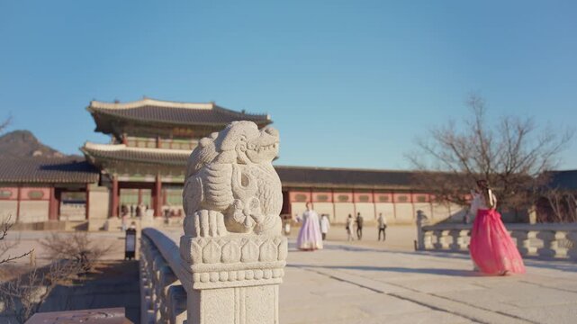Stone haetae guardian statue on ornate bridge at Gyeongbokgung Palace with visitors in colorful traditional hanbok dresses walking through the courtyard under clear blue winter skies in Seoul.