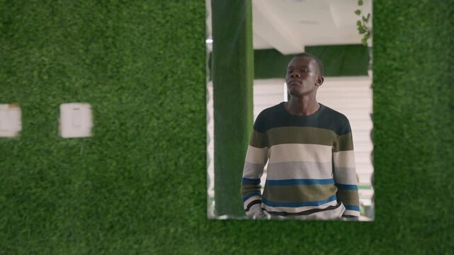 Black man framed by green wall, standing behind rectangular window cutout in bright indoor space, striped sweater, contemplative gaze upward, waiting for date, subtle anticipation, modern minimal