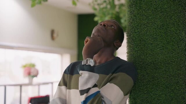 Man waiting in cozy cafe, anxious smile leaning against green wall, adjusting watch, soft sunlight through window, vase of flowers on table, introspective pose conveying romantic anticipation