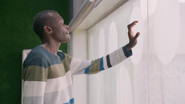 Man touching sheer curtain waiting. Cozy apartment interior with striped sweater, soft daylight filtering through patterned blind, fingertips tracing concentric texture, pensive profile, subtle