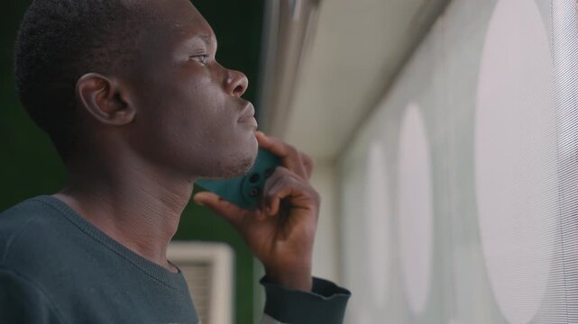 Man holding phone by window waiting, closeup profile of contemplative young man indoors with circular patterned blinds and soft daylight, subtle film grain, tender expression, anxious anticipation