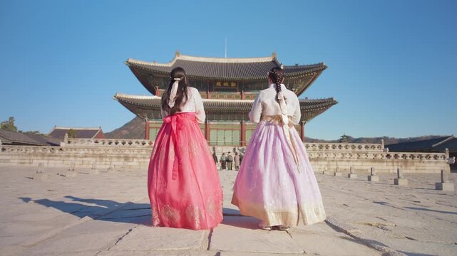Two women in colorful traditional Korean hanbok dresses with pink and lavender skirts stand in the stone courtyard of Gyeongbokgung Palace, gazing at the ornate wooden pavilion under a clear blue sky.