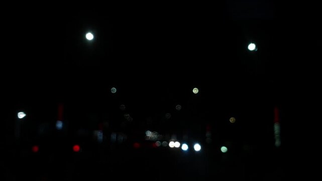 Smooth forward camera motion reveals defocused red and white car lights on a dark urban street. Bright circular bokeh floats dynamically against the black night sky.