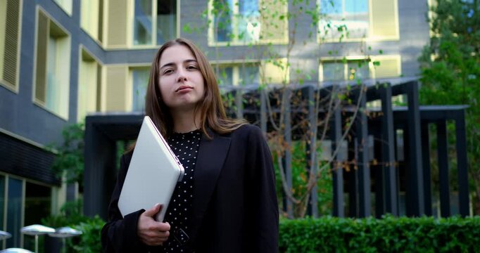 Caucasian woman urban courtyard holding laptop like portfolio, confident pose and decisive hand motion, polka blouse and blazer, strong posture conveying leadership and project ownership
