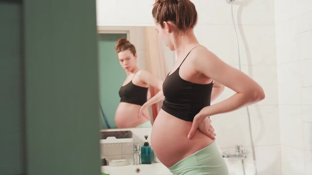 Pregnant caucasian woman inspects belly in bathroom mirror, wearing black crop top and green shorts, hands on lower back and abdomen, gentle massage and posture check, skincare bottles on sink, soft