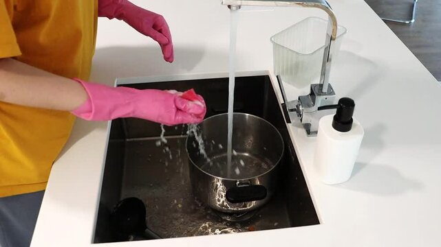 Woman washing cooking pot by hand in kitchen sink with gloves. Home cleaning routine for dish washing, household chores, hygiene, cookware care and domestic lifestyle. Cleaning, big metal sink