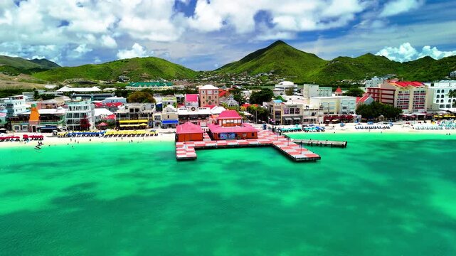 Dynamic aerial drone footage follows a highway road through downtown Philipsburg, St. Maarten. Colorful Caribbean buildings, bustling streets, tropical vegetation, and nearby coastline