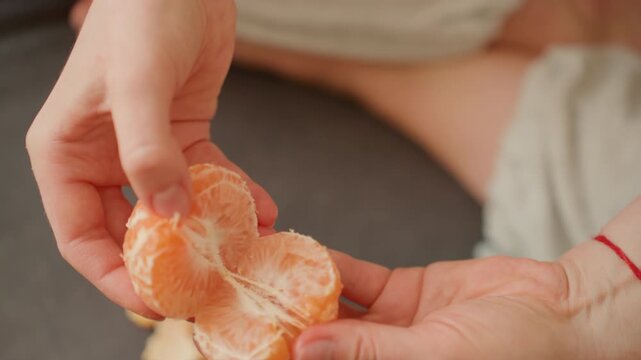 Pregnant woman peeling orange on lap, closeup hands gently separating segments, soft natural light, cozy home setting, expectant mother enjoying vitaminrich snack, red string bracelet visible,