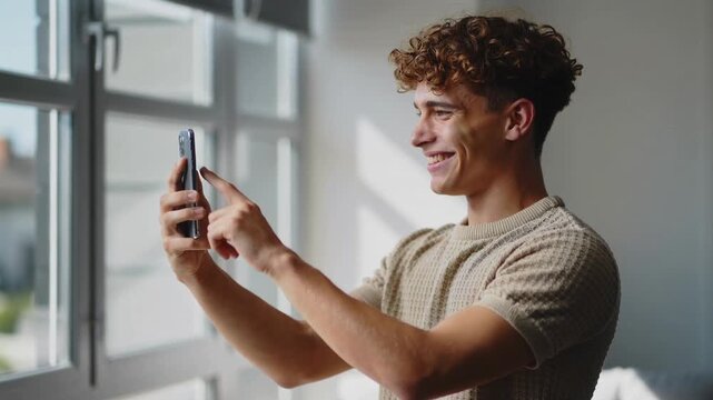 Young man in beige t-shirt using smartphone by window interior medium shot
