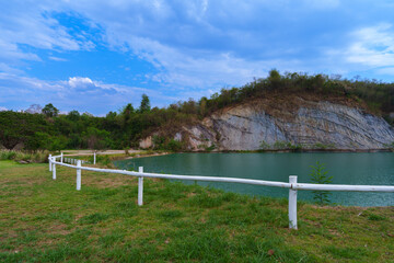 Fototapeta premium Scenic Rural Landscape with White Wooden Fence, Turquoise Pond, and Rocky Cliff under Blue Sky