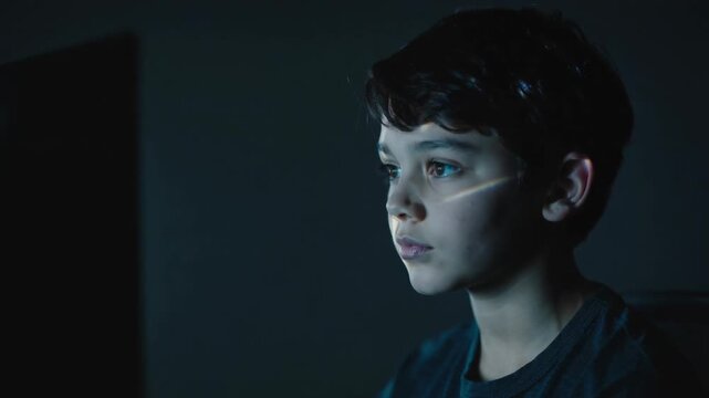 Young boy looking at computer screen in dark room with light reflection, close up