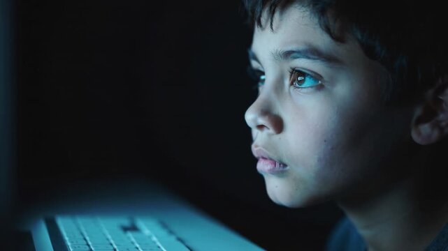 Young boy looking at computer screen in dark room with blue light illumination close up shot