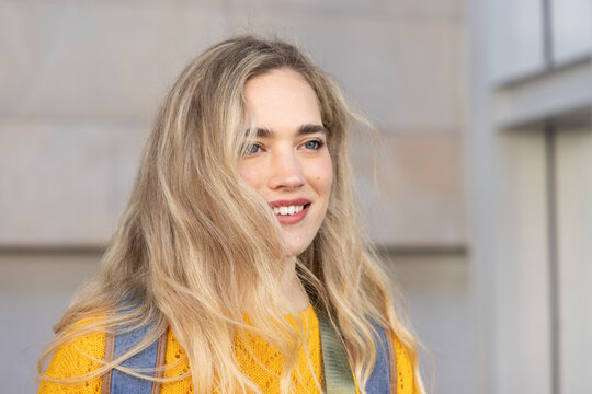 Woman standing by panelled stone wall outdoors, wearing yellow sweater and blue backpack straps