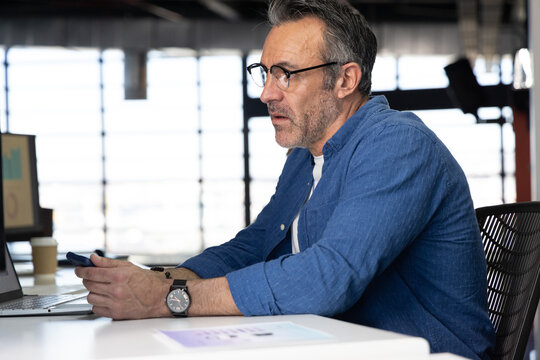 Middle-aged man sitting at white desk in modern office using smartphone and laptop