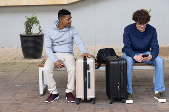Diverse male friends sitting on wooden bench at plaza with beige and black suitcases, smartphone