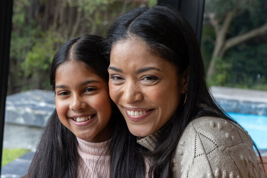 Mother and preteen daughter smiling at home by glass window, in beige and pink knitwear, pool