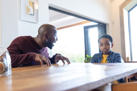 African American father and son sitting at dining table, father gesturing, son looking quiet