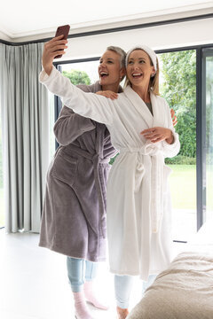 Mother and daughter posing in bright bedroom taking selfie with smartphone wearing plush bathrobes