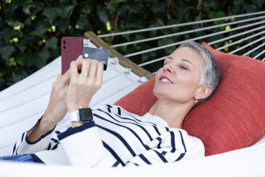 Senior female reclining on hammock in garden, wearing striped top and holding phone and credit card