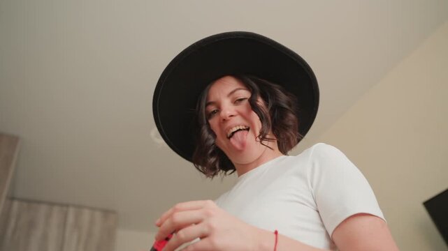 Playful woman in hat indoors low angle portrait showing candid expression sticking out tongue curly hair white tee holding small red object warm soft lighting contemporary room relaxed playful mood