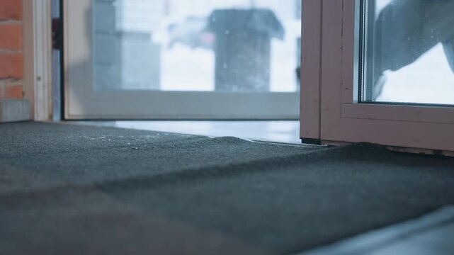 Boots step inside cozy cafe entrance, white student pauses on doormat, leather boots press against mat, snow visible outside through glass door, warm brick interior, quiet winter morning, subtle