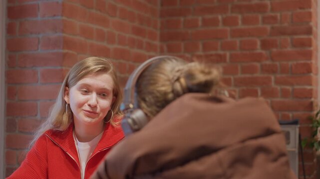Young woman at cafe table, intimate conversation with friend in brown puffer and headphones, exposed brick backdrop, red sweater, attentive glances and small smiles, subtle apology and reassurance,