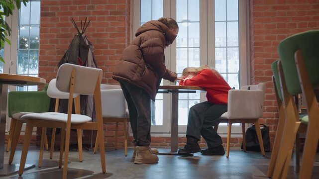 Warm cafe comfort white friend supports after slump. Brick interior and large window, red sweater at table, puffy winter coat stands nearby, gentle hand on shoulder, helping to stand and offer