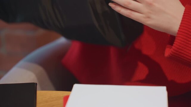 Closeup hands at cafe table, red sweater unpacking notebook and pen, writing notes on spiral pad, focused freelance writer, brick wall background, warm natural light, casual planning moment
