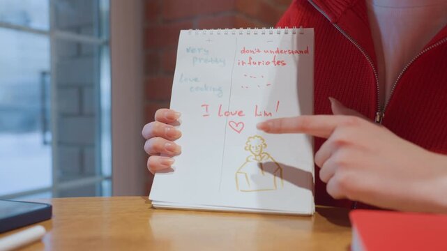 Close up of white hands holding notebook with handwritten love note in cozy cafe, red sweater visible, finger pointing at doodle portrait, textured paper on wooden table by window and brick wall, warm