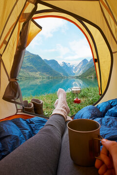 Feet, tent and camping at lake with coffee, holiday and relax for vacation with mountain view. POV, campsite or hand of person with tea outdoor by water for wanderlust on adventure, travel and rest