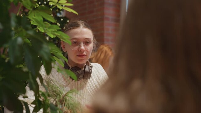 Cozy cafe conversation with white student wearing headphones and glasses, knit sweater, seated by brick wall and lush plants intimate study discussion with soft window light, warm latte, journal notes