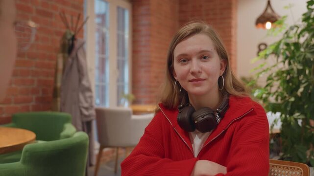 Relaxed cafe conversation with headphones young woman in red sweater sits at table smiling, attentive listener across, warm decor with brick wall and plants, gentle eye contact, nods and laughs,