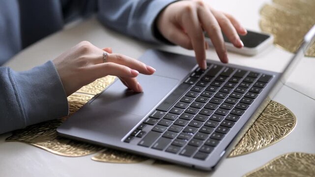 Person uses laptop on golden placemat. Hands type on keyboard and touchpad. Phone lies beside laptop on table. Light shines softly on workspace. Clean and modern working environment