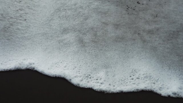 An ocean wave washes over wet black sand, erasing footprints. The mood is calm, serene, and transient, showing the power of nature. Ideal for themes of passage, change, or travel