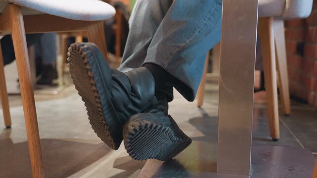 Boots under cafe table closeup showing rubber soles tapping and resting beneath wooden chair in warm urban coffee shop, cropped anonymous patron wearing gray trousers shifts feet, crosses legs, nudges
