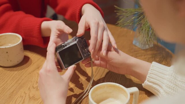 Closeup hands with camera in cafe passing device over latte, red sweater friend reaches to take it, wooden table with mug and small plant, casual playful interaction as they inspect display