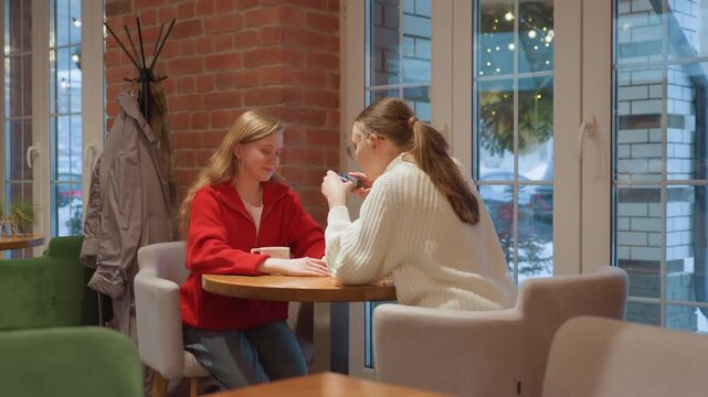 Two white women in cafe photographing heart pose, phone camera capture, cozy knit sweater and red jacket contrast, mugs on table, intimate warm lighting, playful affectionate mood