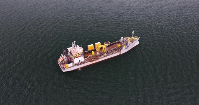 High-quality aerial footage of a dredger support vessel, also known as a subsea cable ship, operating in a busy industrial port. The vessel is designed for underwater cable installation, maintenance, 