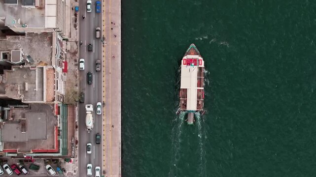 Cinematic aerial top-down tracking shot following a vessel cruising along the coastline. On one side, busy city streets with moving cars and rooftops reflect urban life, while on the other side