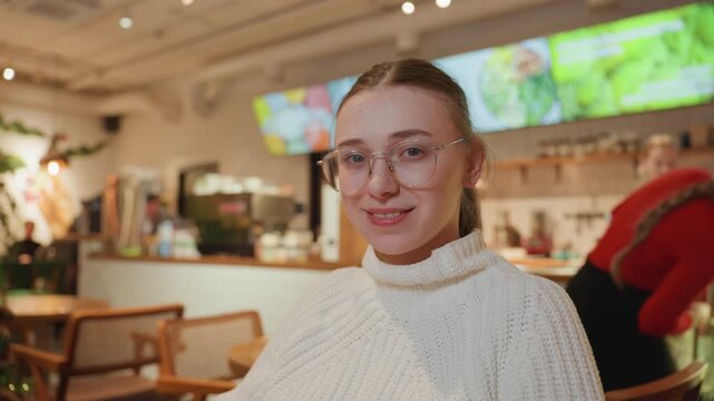 Young white woman in cafe wearing chunky knit sweater and round glasses, seated near decorated tree with bokeh lights, barista in red uniform behind counter, digital menu screens visible, warm golden