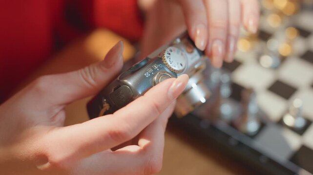 Hands tuning camera on cafe chessboard. Warm amber light, red sweater sleeves, wooden table with chess pieces blurred, compact camera dial being adjusted, deliberate slow movements, thoughtful